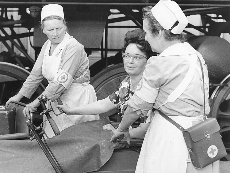 DRK der DDR: Bahnhofsdienst. Hilfe beim Umsteigen auf dem Bahnsteig für eine Frau im Rollstuhl, vermutlich in Dresden